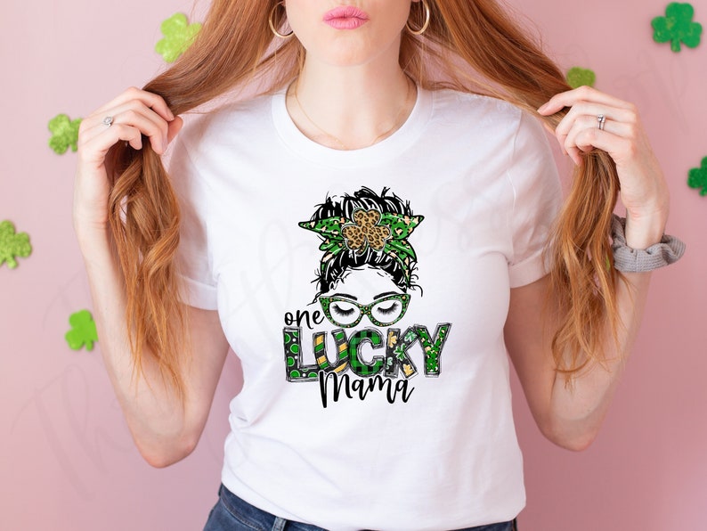 May include: White t-shirt with a green, gold, and black graphic of a woman with a shamrock headband and the text "one lucky mama".