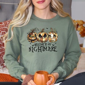 Op de afbeelding: Olijfgroene sweatshirt met een Halloween-ontwerp. Het heeft drie patchwork pompoenen met enge gezichten en de tekst "WELCOME TO MY NIGHTMARE" eronder. Het ontwerp bevat vleermuizen en decoratieve krullen, perfect voor de herfst.