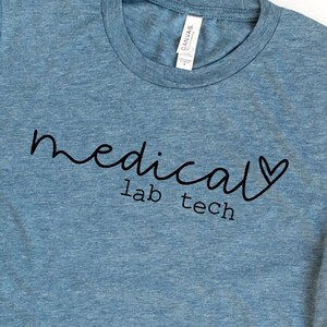 May include: A light blue t-shirt with the words "medical lab tech" in black script. The word "medical" is written in a cursive font, and a heart replaces the letter "a" in the word. The shirt has a crew neck.