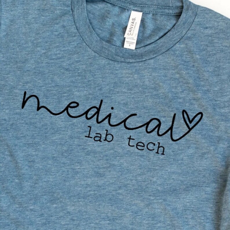 May include: A light blue t-shirt with the words "medical lab tech" in black script. The word "medical" is written in a cursive font, and a heart replaces the letter "a" in the word. The shirt has a crew neck.