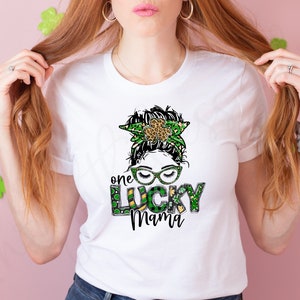 May include: White t-shirt with a green, gold, and black graphic of a woman with a shamrock headband and the text "one lucky mama".