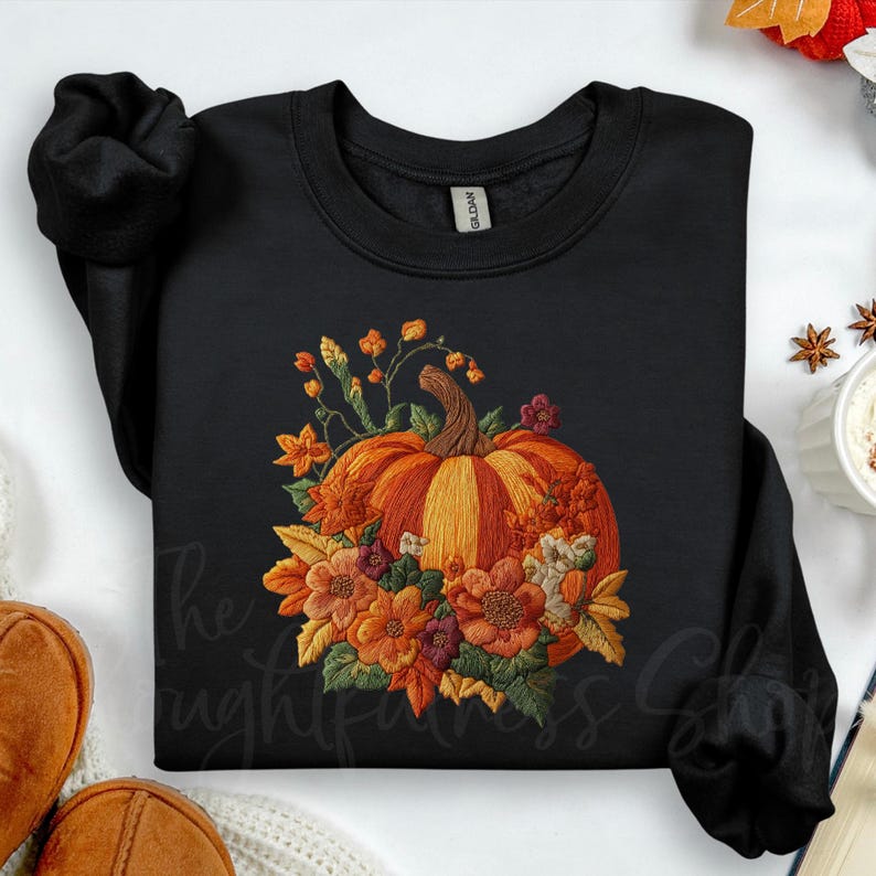 A black sweatshirt with a vibrant pumpkin embroidered on it, surrounded by autumn flowers, is displayed on a white background.