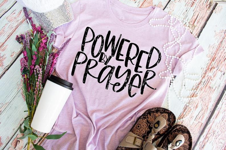 May include: A pink t-shirt with the text "Powered by Prayer" in black distressed lettering. The shirt is laid flat on a wooden surface with a white mug, a string of pearls, and a pair of sandals.