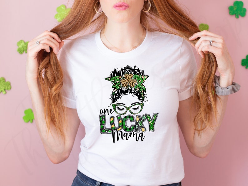 May include: White t-shirt with a green, gold, and black graphic of a woman with a shamrock headband and the text "one lucky mama".
