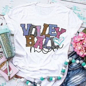 Pu&ograve; includere: Maglietta bianca con le parole "Volleyball Mom" in lettere colorate con un design a forma di cuore. Le lettere sono decorate con strisce, stampa leopardata e altri motivi.