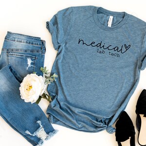 May include: A blue t-shirt with the words "medical lab tech" in black script. The shirt is paired with distressed blue jeans, a white flower, and black flats. The items are arranged on a white surface.