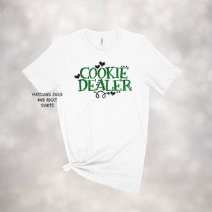 Puede incluir: Camiseta blanca con texto en purpurina verde que dice "Cookie Dealer" con corazones en purpurina negra. El texto tambi&eacute;n dice "Matching Child and Adult Shirts".
