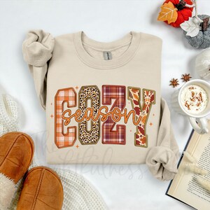 Può includere: Una felpa beige con la scritta "COZY SEASON" in una miscela di caratteri a fantasia, tra cui scozzese e stampa leopardata. La felpa è piegata, con una tazza di caffè, un libro e decorazioni a tema autunnale sullo sfondo.