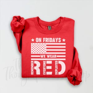 Könnte beinhalten: Ein rotes Sweatshirt mit dem Text "ON FRIDAYS WE WEAR RED" und einem Design der amerikanischen Flagge. Die Worte "REMEMBER EVERYONE DEPLOYED" sind vertikal auf beiden Seiten des Wortes "RED" aufgedruckt.