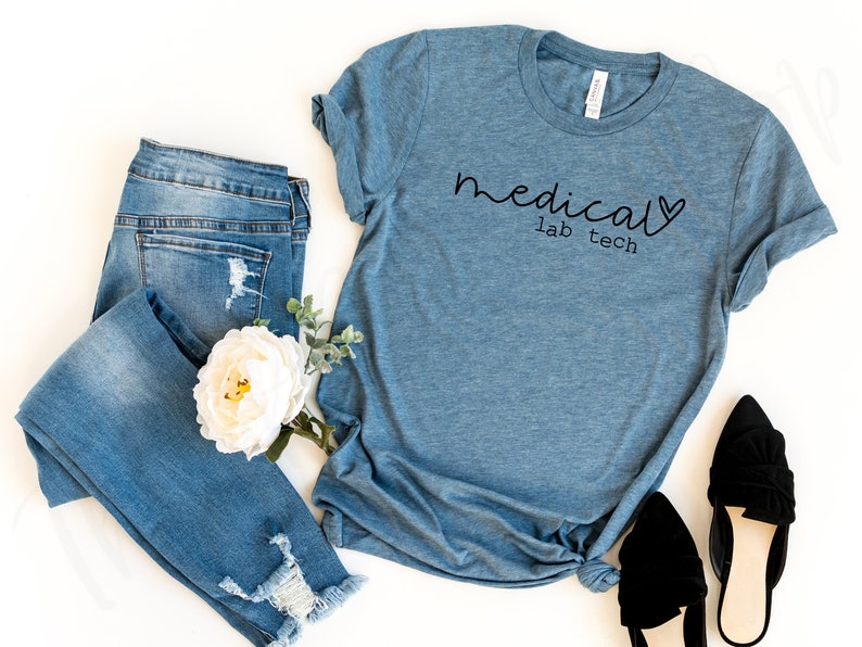 May include: A blue t-shirt with the words "medical lab tech" in black script. The shirt is paired with distressed blue jeans, a white flower, and black flats. The shirt is made of a soft material.