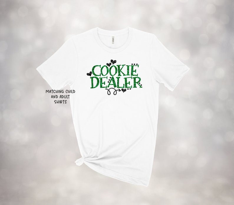 Puede incluir: Camiseta blanca con texto en purpurina verde que dice "Cookie Dealer" con corazones en purpurina negra. El texto tambi&eacute;n dice "Matching Child and Adult Shirts".