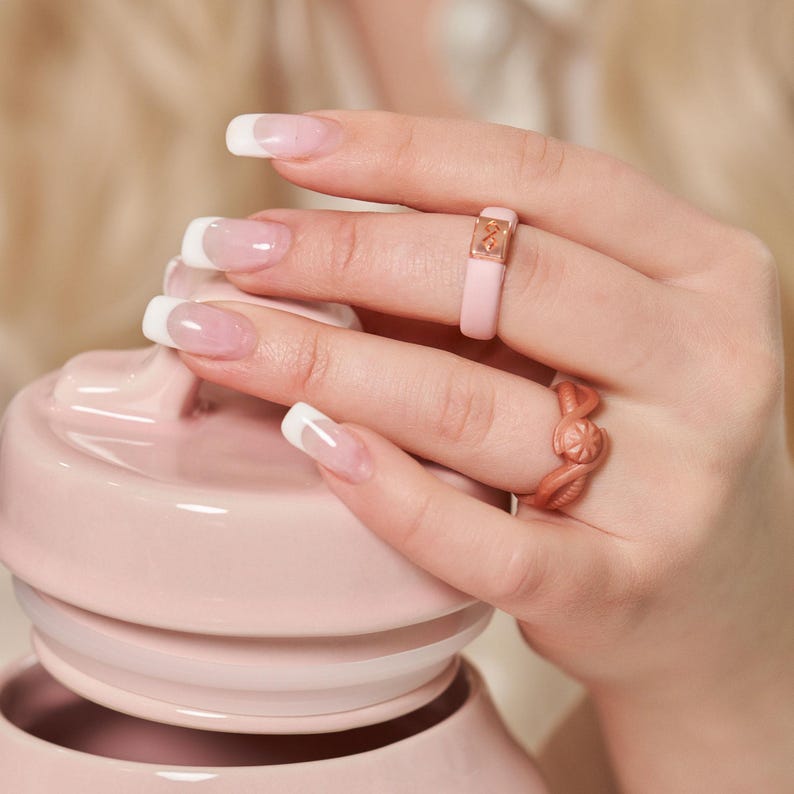 Op de afbeelding: Twee siliconen ringen op een hand. De ring op de wijsvinger is roze met een gouden accent. De ring op de middelvinger is een gedraaid ontwerp in een roze gouden kleur.