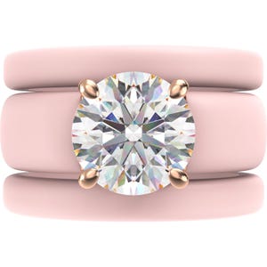 May include: A three-piece ring set featuring a large, round-cut diamond set in a gold-colored prong setting. The diamond is the focal point, surrounded by two light pink bands. The bands have a smooth, rounded design.