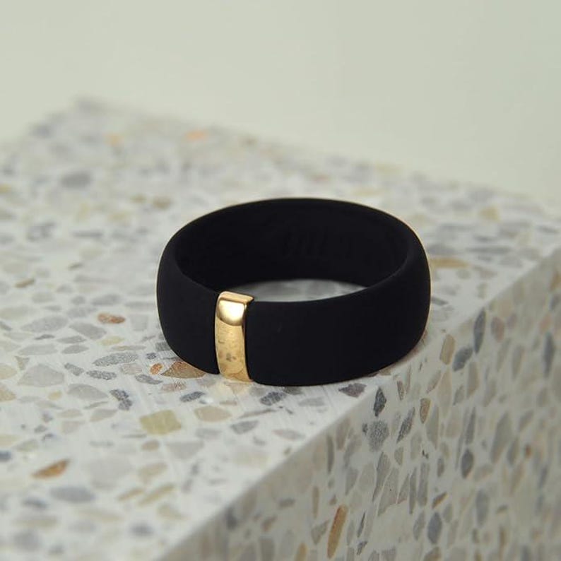 May include: A black silicone ring with a gold band. The ring is on a white and gray speckled surface.