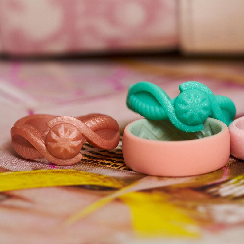 Op de afbeelding: Twee siliconen ringen met een swirl-ontwerp en een stervormig accent. De ene ring is lichtroze en de andere is turquoise.