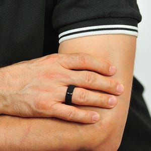 May include: A black silicone ring with a black inlay. The ring is worn on the ring finger of a person's left hand.