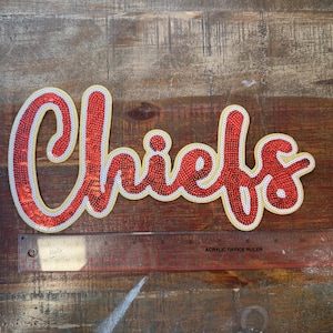 May include: A red sequined patch featuring the word Chiefs in a cursive script with a white and yellow border. A clear plastic ruler is positioned underneath, showing the patch measures approximately 11 inches in total length.