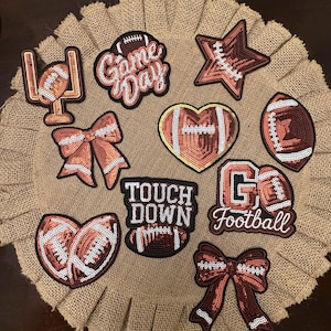 May include: A collection of football-themed iron-on patches featuring sequins in shades of brown, white, and black. Designs include a goalpost, a bow, a heart-shaped football, a star, and text reading Game Day, TOUCH DOWN, and Go Football.