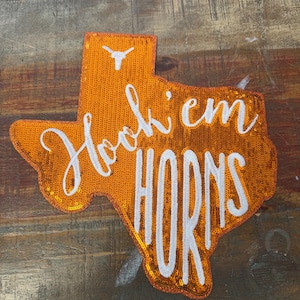 May include: A Texas-shaped patch covered in orange sequins featuring the white embroidered text Hook 'em HORNS and a small white longhorn steer logo at the top. The patch is designed for application to fabric items and displays a vibrant, reflective surface.