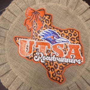 May include: A circular burlap wall hanging featuring a sequined patch shaped like the state of Texas. The patch has a leopard print pattern, an orange bow, the text UTSA in orange, and the word Roadrunners in white with a blue bird mascot.