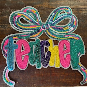 May include: A decorative sign featuring the word teacher in colorful, sequined letters. The letters are pink, green, purple, and yellow, outlined in silver. Above the text is a large, matching sequined bow in teal, pink, and yellow stripes.