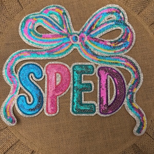 May include: A decorative sign featuring the word SPED in large, sequined letters. The letter S is blue, P is pink, E is teal, and D is purple. Above the text is a large, multi-colored sequined bow with silver trim, all set against a round burlap background.