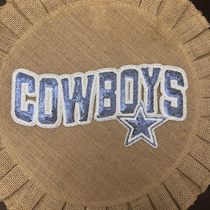 May include: A circular tan burlap mat with a pleated border featuring a large, blue and silver sequined patch that spells out the word COWBOYS in capital letters, accompanied by a matching blue and silver sequined star emblem positioned below.