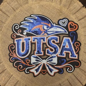 May include: A circular burlap wall hanging featuring a sequined UTSA logo in blue and orange. The design includes a bird mascot head, the text UTSA, a decorative bow, and heart accents, all outlined in black against the natural tan burlap background.