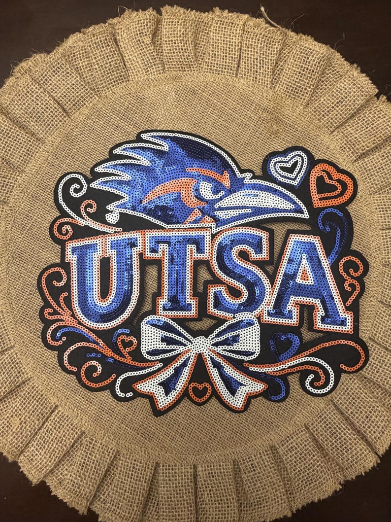 May include: A circular burlap wall hanging featuring a sequined UTSA logo in blue and orange. The design includes a bird mascot head, the text UTSA, a decorative bow, and heart accents, all outlined in black against the natural tan burlap background.