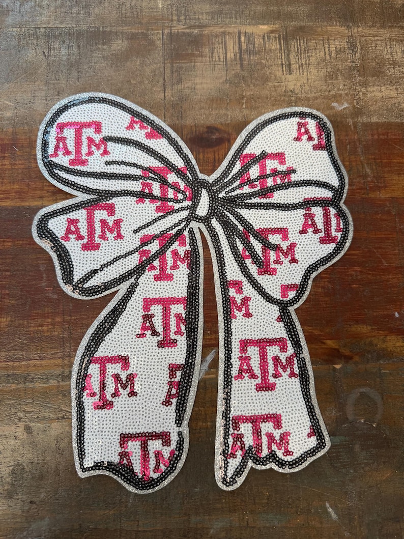 May include: A decorative bow patch featuring a white background covered in a repeating pattern of the maroon Texas A&M University ATM logo. The entire surface is embellished with small, shimmering sequins, and the edges are outlined in black.