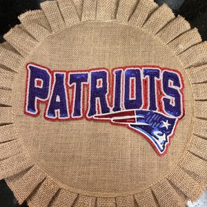May include: A circular tan burlap wall hanging featuring a pleated border. The center displays the word PATRIOTS in blue sequined lettering with a red outline, accompanied by a blue, red, and white Patriots team logo patch on the right side.