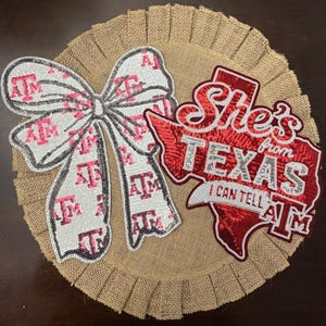 May include: A circular burlap wall hanging featuring a white sequined bow with a repeating maroon ATM logo. Next to the bow is a red sequined cutout of the state of Texas with the text She's from TEXAS I CAN TELL ATM in white sequins.