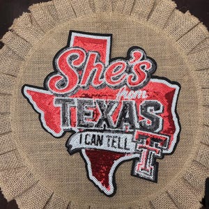 May include: A circular burlap wall hanging featuring a red sequined map of Texas. The text reads She's from TEXAS I CAN TELL with a Texas Tech logo. The burlap has a pleated, ruffled edge, and the lettering is outlined in black and white.