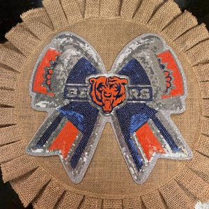 May include: A decorative bow featuring a sequined Chicago Bears logo in navy blue, orange, and silver. The bow is centered on a circular, pleated tan burlap base. The word BEARS is visible in the center of the bow, accompanied by a bear head icon.