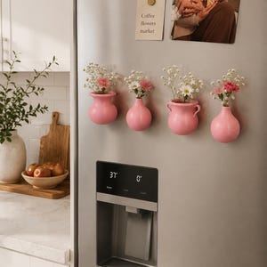 May include: Four small, matte pink ceramic vases attached to a stainless steel refrigerator. Each vase holds a miniature bouquet of white baby's breath and small pink or white flowers. The vases have varying rounded shapes and one features side handles.