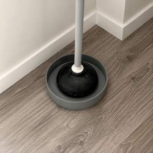 Op de afbeelding: Een zwarte rubberen ontstopper met een witte kraag en een lange lichtgrijze steel, rustend in een ronde donkergrijze plastic lekbak. De ontstopper heeft een geribbelde koepelvorm aan de basis, ontworpen voor het ontstoppen van afvoeren.