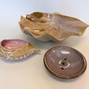 May include: A collection of three ceramic items: a large, tan, shell-shaped bowl with a glossy finish, a smaller, tan, crab-shaped dish with a smooth pink interior, and a round, speckled, mauve-colored soap dish with three drainage holes.