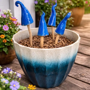 May include: A ceramic planter with a blue and white ombre design holds several blue mushroom garden stakes. The stakes have a glossy finish and are placed in a bed of mulch. The planter sits on a wooden deck.