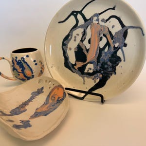 May include: A ceramic set consisting of a round plate, a mug, and a small bowl. Each piece is off-white with a marbled design featuring swirls of dark blue, light blue, and peach paint. The plate is held upright by a black metal stand.