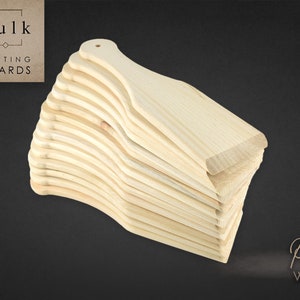 May include: A stack of 15 unfinished wooden chopping boards made of pine wood. The boards are all the same size and shape, with a rounded handle on one end. The boards are stacked in a staggered pattern, with the top board slightly offset from the bottom board. The boards are all a light brown colour.