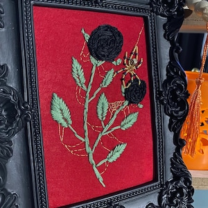 May include: Embroidered artwork in a black ornate frame. The red fabric background features a black rose and a spider with gold accents. Green leaves and gold thread spiderwebs add detail to the floral design.