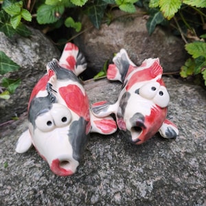 Könnte beinhalten: Zwei Koi-Fisch-Figuren aus Keramik mit weißem Grund und Flecken in Rot und Dunkelgrau. Jeder Fisch hat große, hervorstehende Augen und ein offenes, kreisförmiges Maul. Die Oberfläche der Keramik ist glatt und glänzend gestaltet.