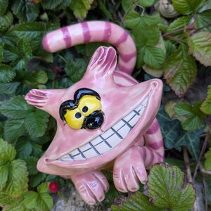 Könnte beinhalten: Eine rosa Keramikstatue der Grinsekatze mit breitem Grinsen, gelben Augen und schwarzen Pupillen. Die Katze hat einen gestreiften Schwanz und ist von grünen Blättern umgeben. Eine skurrile Gartendekoration.