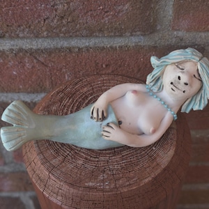 Könnte beinhalten: Keramik-Meerjungfrau-Figur mit blauen Haaren und türkisfarbenem Schwanz, ruhend auf einem Holzpfosten. Die Meerjungfrau trägt eine blaue Perlenkette. Der Hintergrund ist eine rote Backsteinmauer.