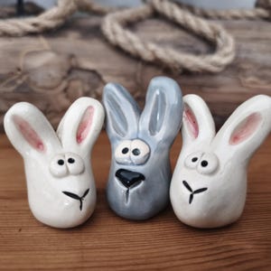 Könnte beinhalten: Drei Keramik-Hasenfiguren. Zwei sind weiß mit rosa gefütterten Ohren, und einer ist grau. Jede hat ein schwarzes, stilisiertes Gesicht mit einer kleinen, schwarzen Nase und einem schwarzen, umgedrehten Y-förmigen Mund. Die Hasen stehen auf einer Holzoberfläche.