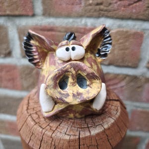 Könnte beinhalten: Eine Keramikskulptur eines Wildschweinkopfes mit einer gesprenkelten braunen und gelben Glasur. Die Figur hat große weiße Augen mit schwarzen Pupillen, markante weiße Stoßzähne und dunkle, strukturierte Ohren. Die Schnauze ist breit.