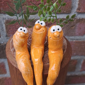 Könnte beinhalten: Drei orangefarbene Keramik-Karottenfiguren mit glänzender Oberfläche, jede mit einem lächelnden Gesicht mit weißen Augen und schwarzen Pupillen, gekrönt von grünem Kunststofflaub. Die Karotten haben strukturierte Oberflächen.