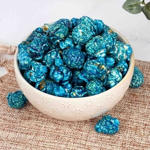 May include: A white bowl filled with blue popcorn. The popcorn is a light blue color with some white kernels. The bowl is sitting on a brown woven surface.