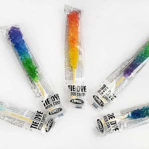 May include: An overhead shot of six individually wrapped rock candy sticks. Each stick is a different color gradient, including rainbow, blue, green, and orange. The packaging reads "TIE DYE ROCK CANDY".