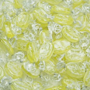 May include: A close-up of a pile of yellow lemon flavored candies wrapped in clear cellophane. The candies are oval shaped and have a white logo on them.